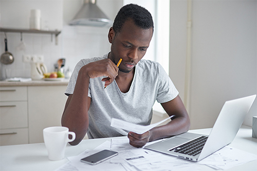 Young man sitting in a kitchen, looking at bills with a computer and cup of coffee on a table. 