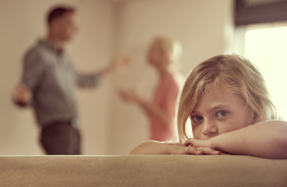 A young child at home watching her parents fight and argue about money behind her.